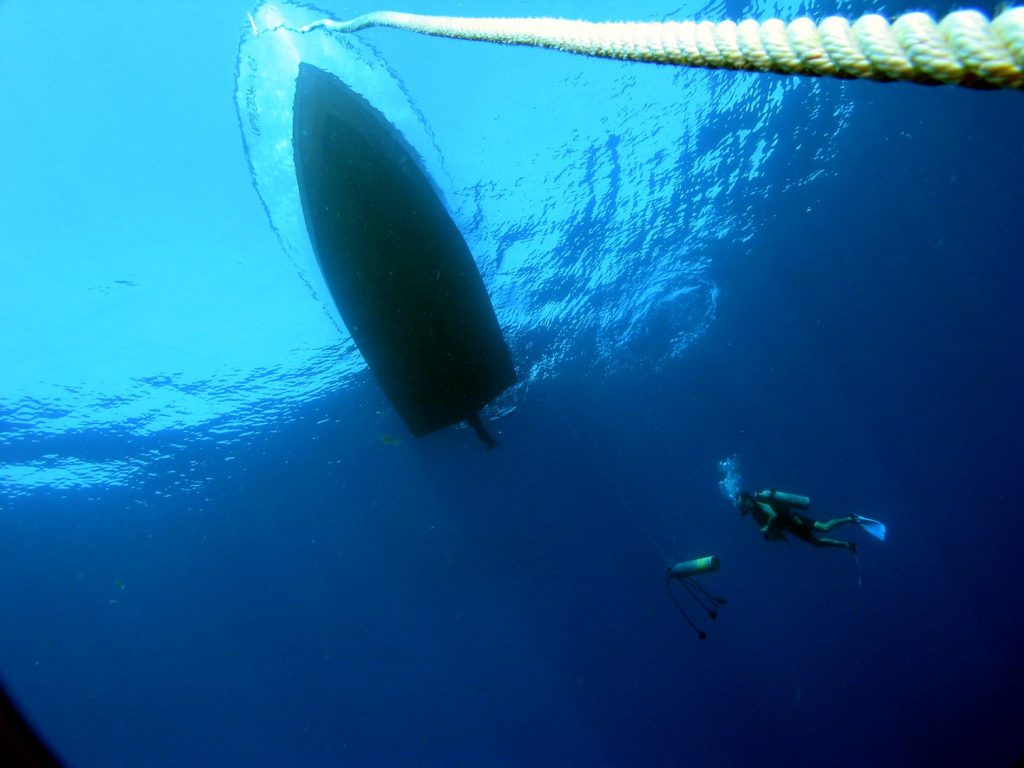 Underwater photo with the view of the shadowed dark bottom of a boat, the anchor roap and a lone diver approaching the boat. 