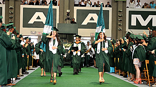 Photo of UPRM graduation. Two graduate students bear UPRM flags as they march  during the ceremony.