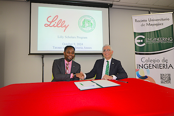 Sentados en una mesa desde la izquierda, Brandon Paschal, vicepresidente senior de Lilly del Caribe, Inc. y el doctor Agustín Rullán Toro, rector del RUM, se saludan tras el acuerdo del programa de becas. Atrás proyectados en una pantalla los logos de Lilly y el RUM.