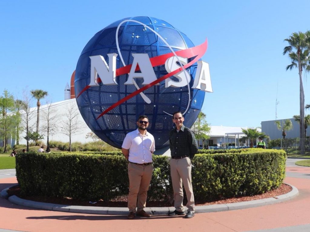 Los investigadores principales, de pie, frente al globo con el símbolo de NASA en las instalaciones del Centro Espacial John F. Kennedy en Cabo Cañaveral.